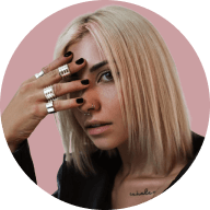 Blonde woman with straight hair covering part of her face with her hand wearing multiple silver rings, against a muted pink circular background.